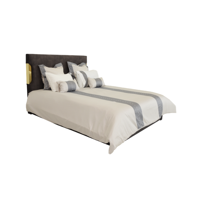 Cama com cabeceira estofada cinzenta escura e detalhe dourado. Edredão bege com duas faixas cinzentas pontilhadas e almofadas a condizer.