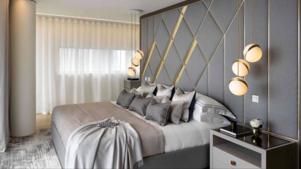 Quarto moderno com cama grande, cabeceira estofada cinza com padrão geométrico e detalhes dourados, cortinas brancas e candeeiros pendentes esféricos.