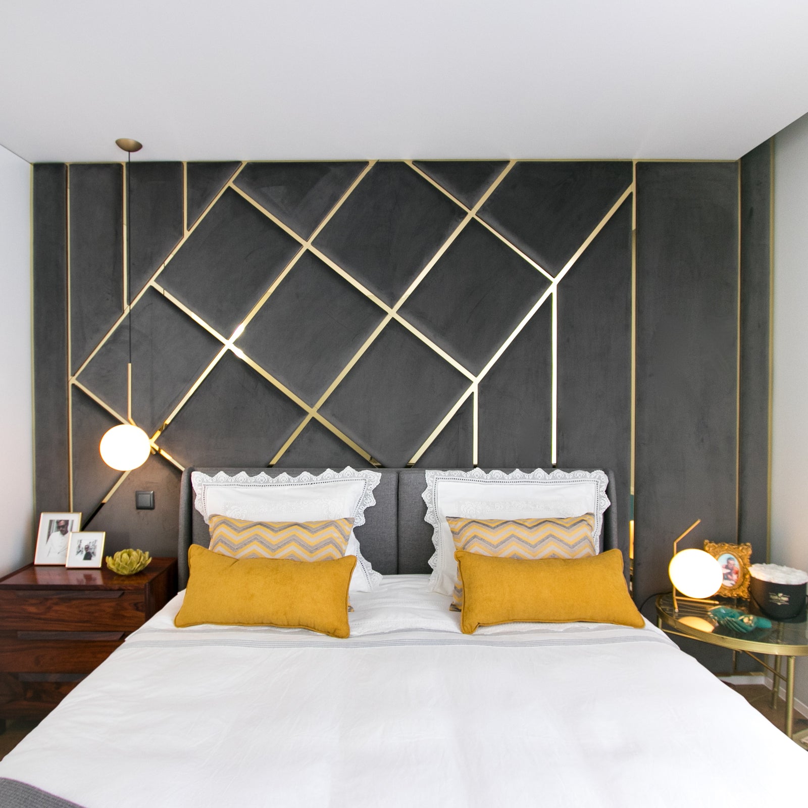 Quarto moderno com cama de casal, roupa de cama branca e almofadas amarelas. Painel de parede estofado cinzento com padrão geométrico dourado.