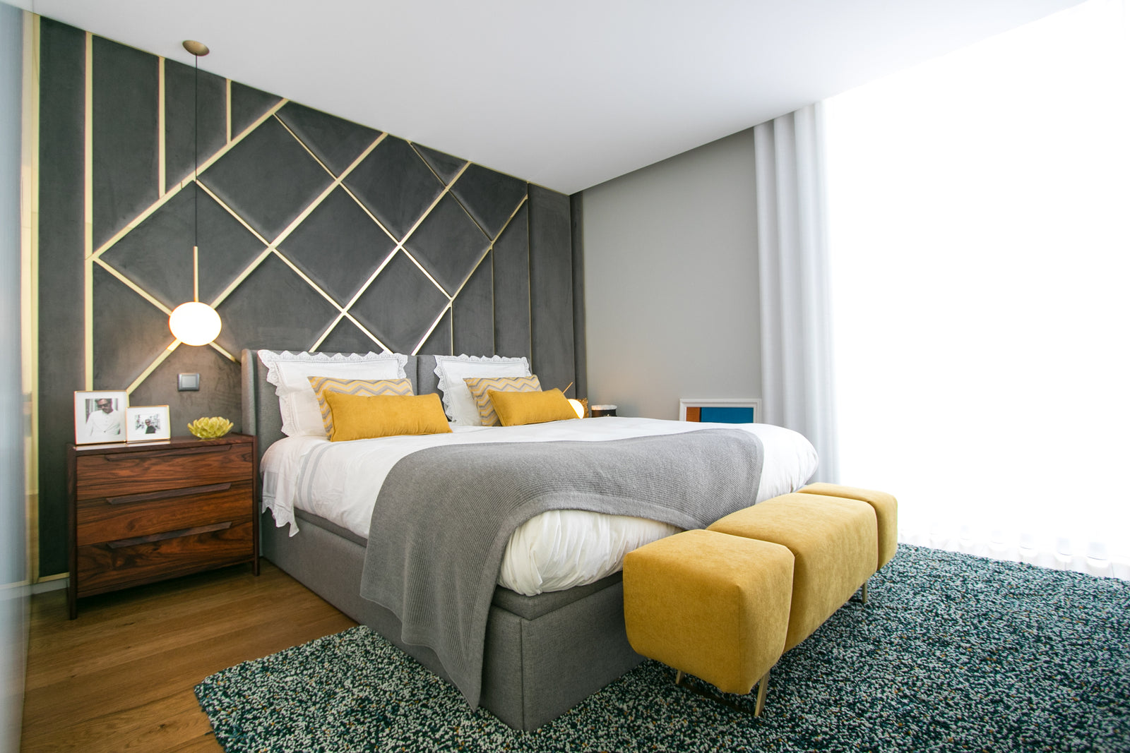 Quarto moderno com cama de casal, cabeceira cinzenta com padrões dourados, almofadas amarelas, duas banquetas amarelas e tapete verde.