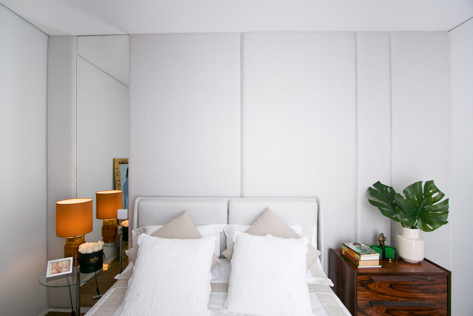 Quarto de casal com cabeceira e parede estofadas cinza. Mesas de cabeceira com candeeiros laranja, livros e planta Monstera.
