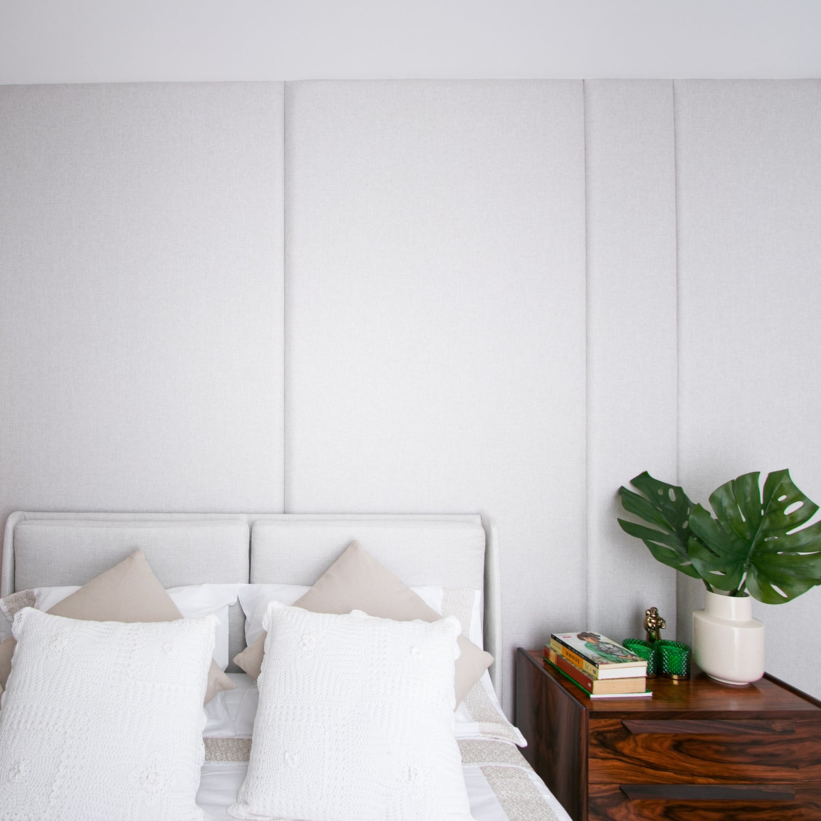 Quarto moderno com cama de casal, cabeceira estofada cinza, almofadas brancas e bege, e mesa de cabeceira de madeira escura com livros e planta.