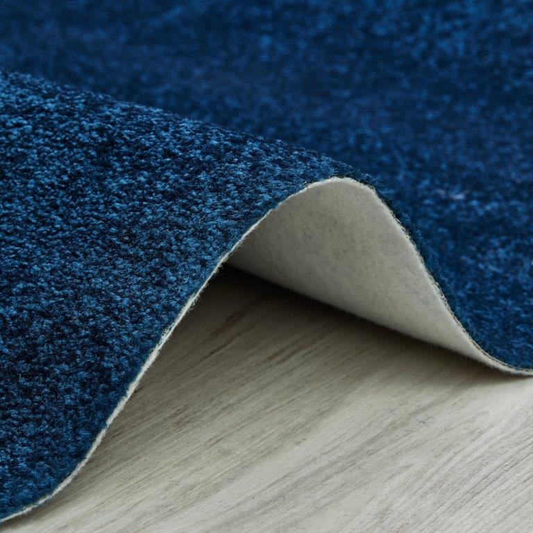 Tecido azul escuro com textura, parcialmente enrolado, revelando o seu verso claro sobre um pavimento de madeira.