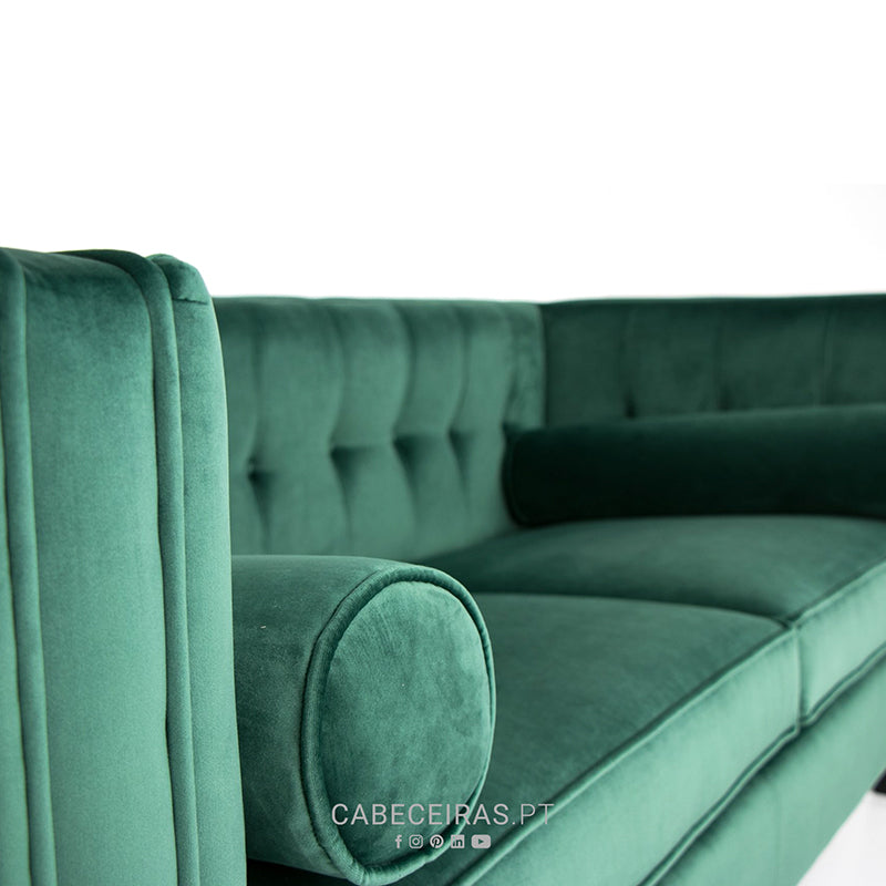 Sofá de veludo verde esmeralda com encosto capitonê e almofada cilíndrica, em fundo branco.