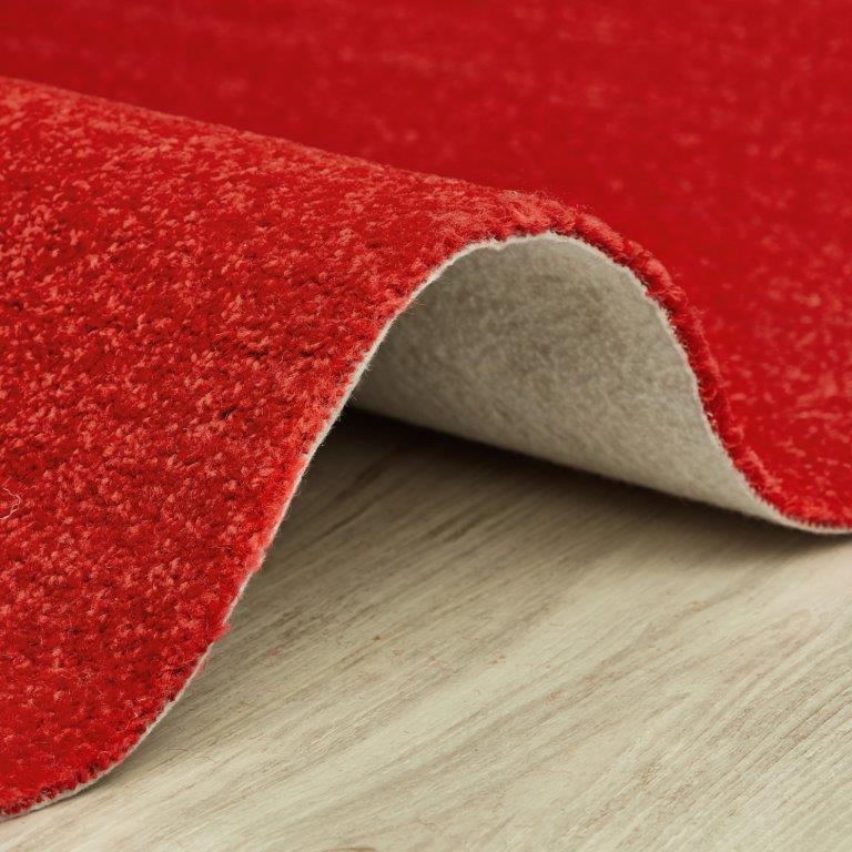 Tapete vermelho enrolado, mostrando a superfície texturada, o verso cinzento e o chão de madeira clara.
