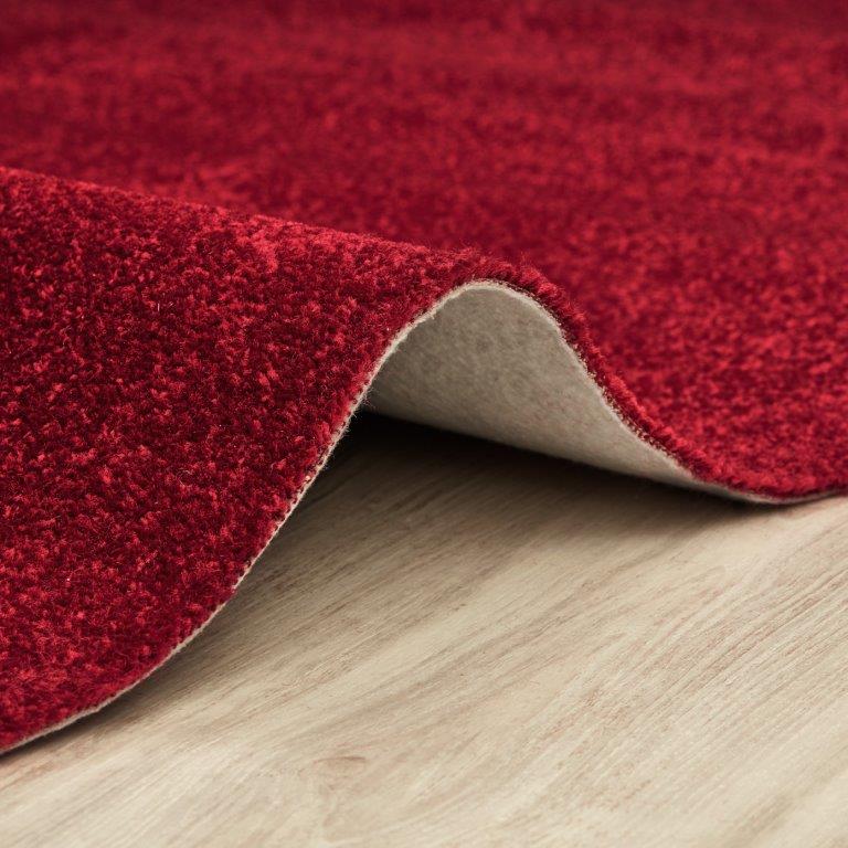 Tapete vermelho com textura visível, levantado para mostrar o verso bege sobre um chão de madeira clara.